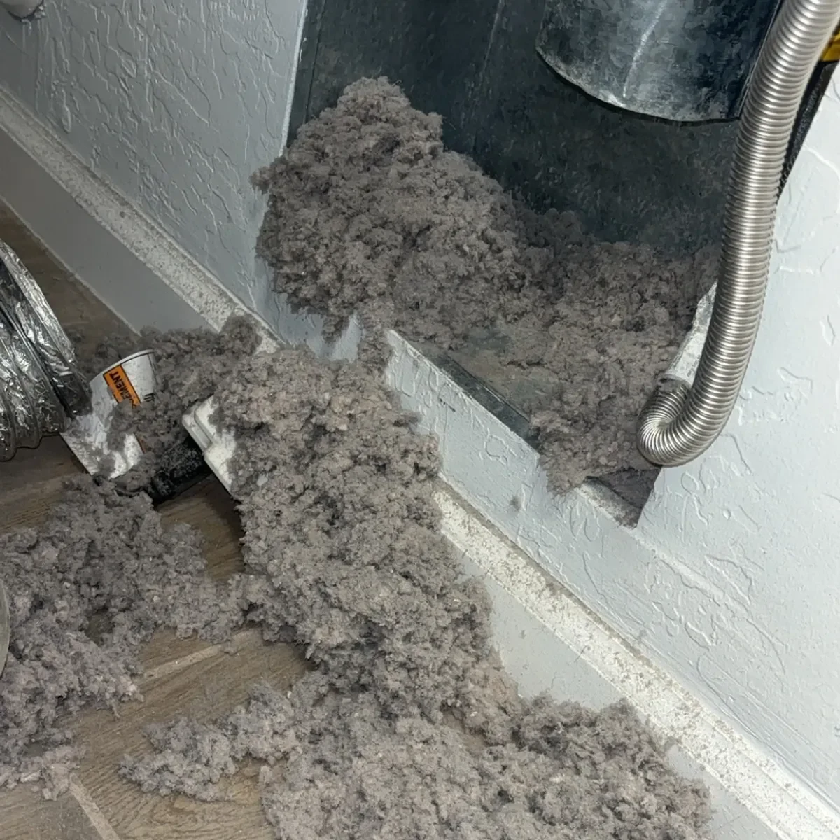 Enormous pile of gray dryer lint spilling out of a wall vent opening onto a laundry room floor