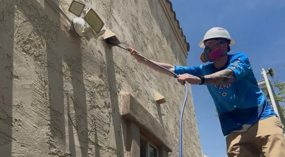 Technician in hard hat and respirator running a cleaning line into an exterior dryer vent on a stucco wall